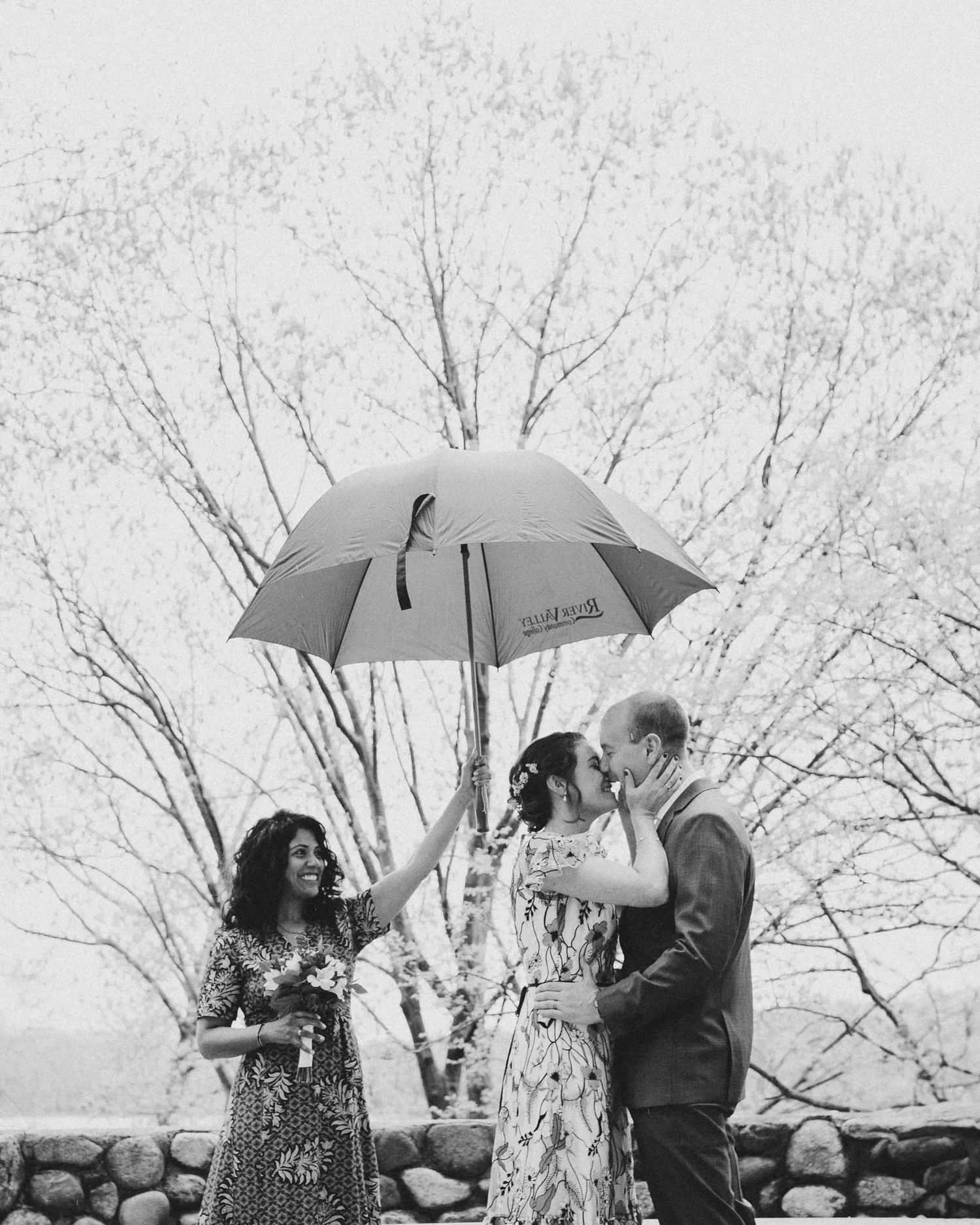 A black and white photo of a couple kissing under an umbrella with a woman next to them holding a bouquet, outdoors with a stone wall and trees in the background during a Boston elopement.