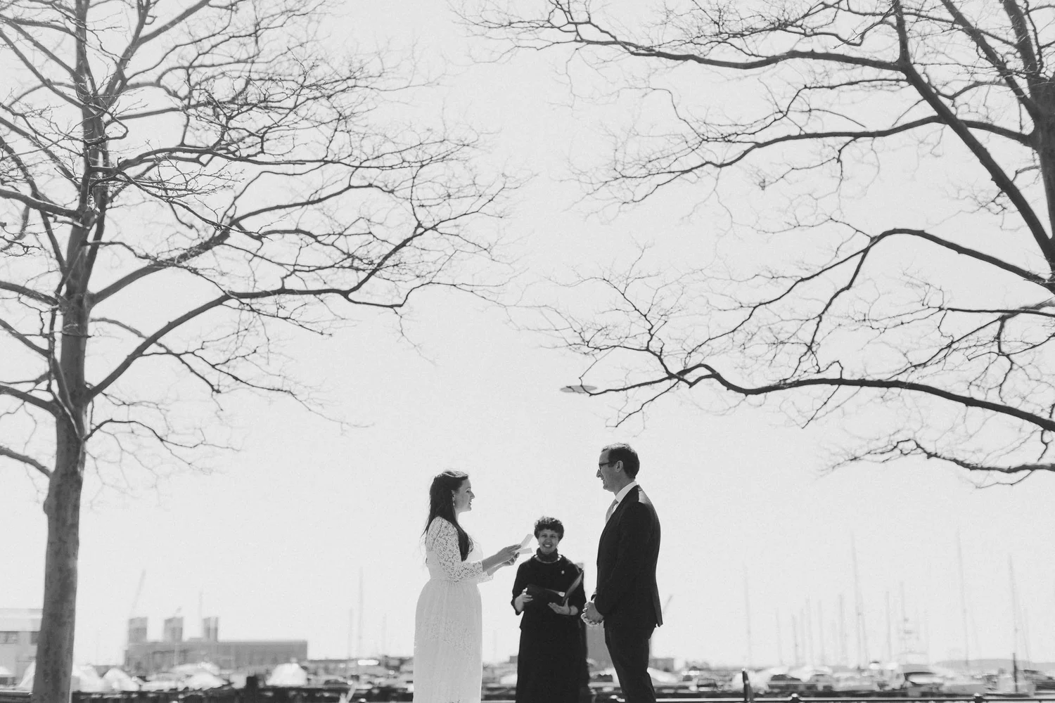 A couple in wedding attire exchanging vows in an outdoor Boston elopement wedding ceremony near a marina with boats and sailboats, with leafless trees in the background.