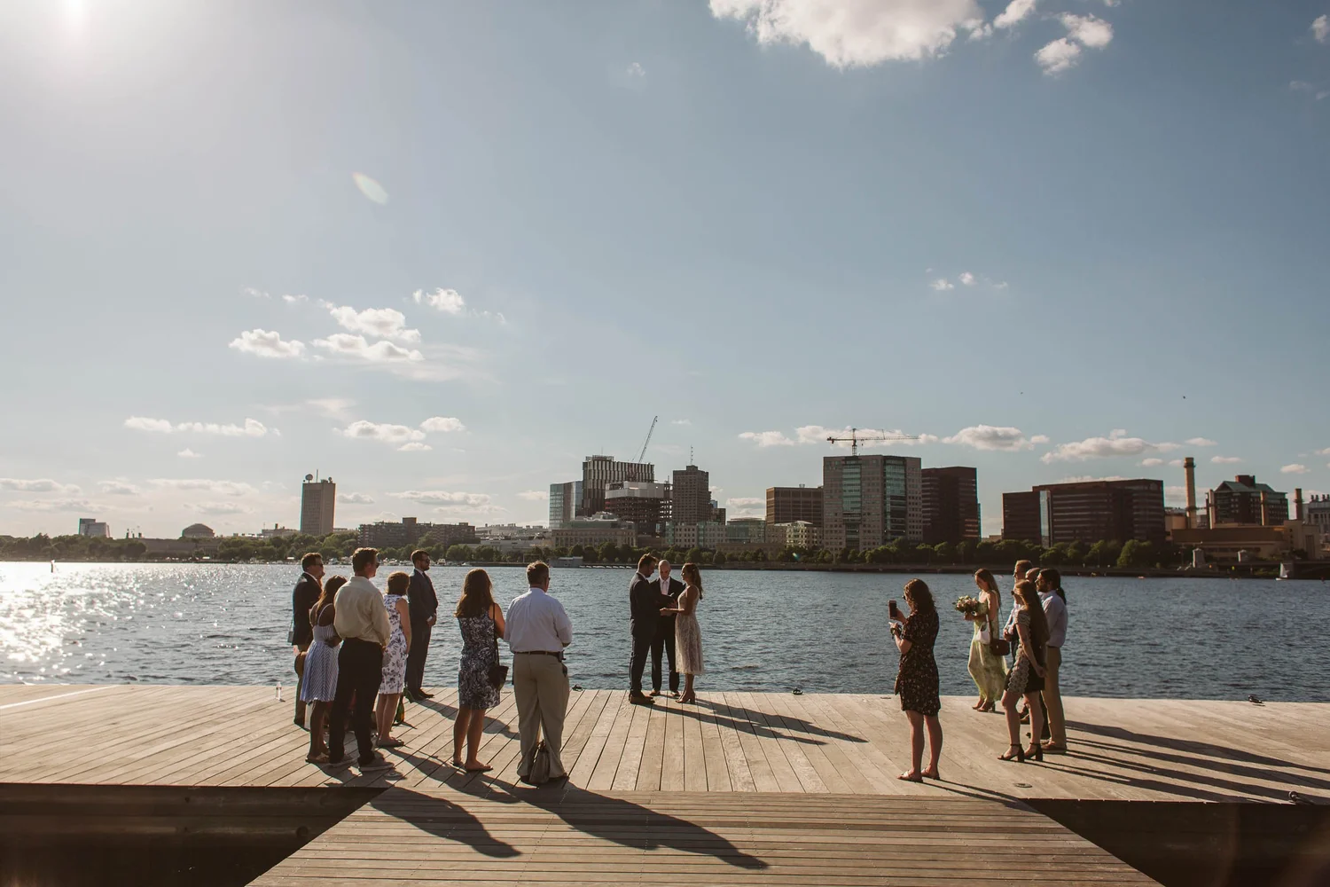 A elopement wedding ceremony taking place on a wooden dock along the Charles River in Boston with the Cambridge skyline in the backgroundwith people gathered around, some taking photos, during sunset.