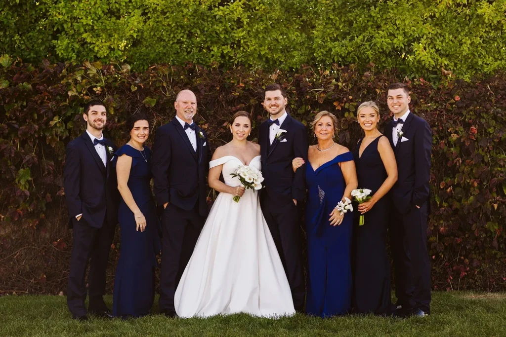Bride, groom and wedding party in navy stand in front of the tall green hedge at Wychmere Beach Club for a group photo
