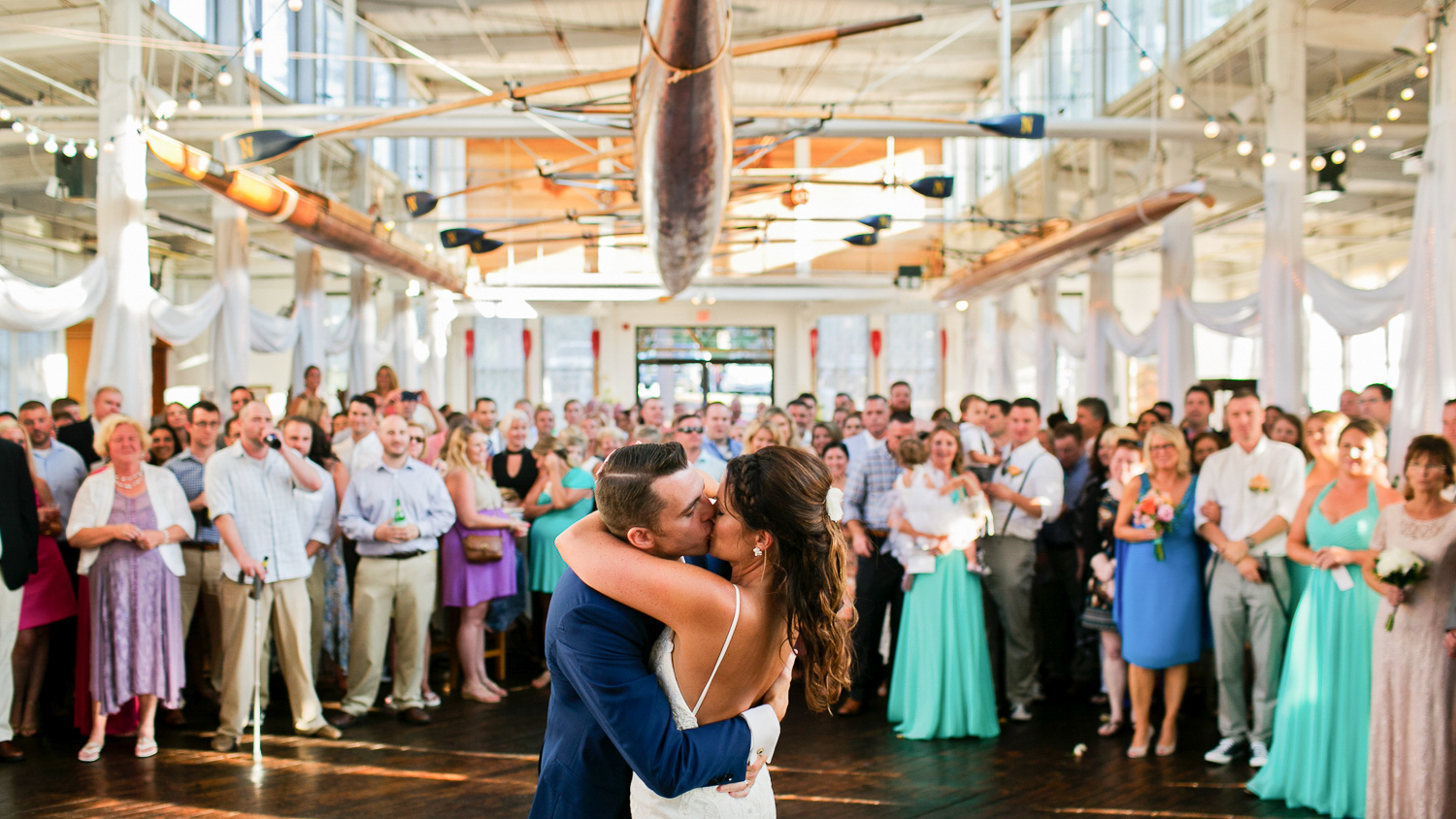 Courageous Sailing Center Wedding Information and Guide 1 Couple kiss on dancefloor surrounded by crowd beneath hanging rowing shells