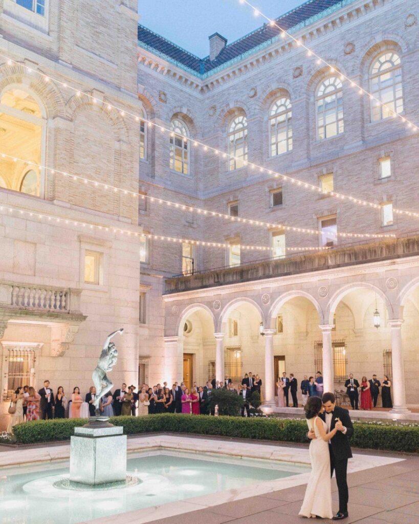 Couple dancing alone in the Boston Public Library courtyard at dusk, guests watching from the arcade beneath strings of cafe lights
