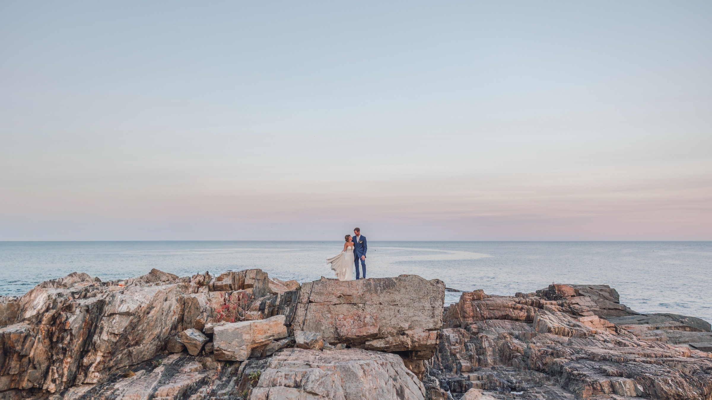 Team Lead Photographer Eric 1 Bride and groom standing together on the rocks above the ocean at dusk on the New England coast, photographed by Eric