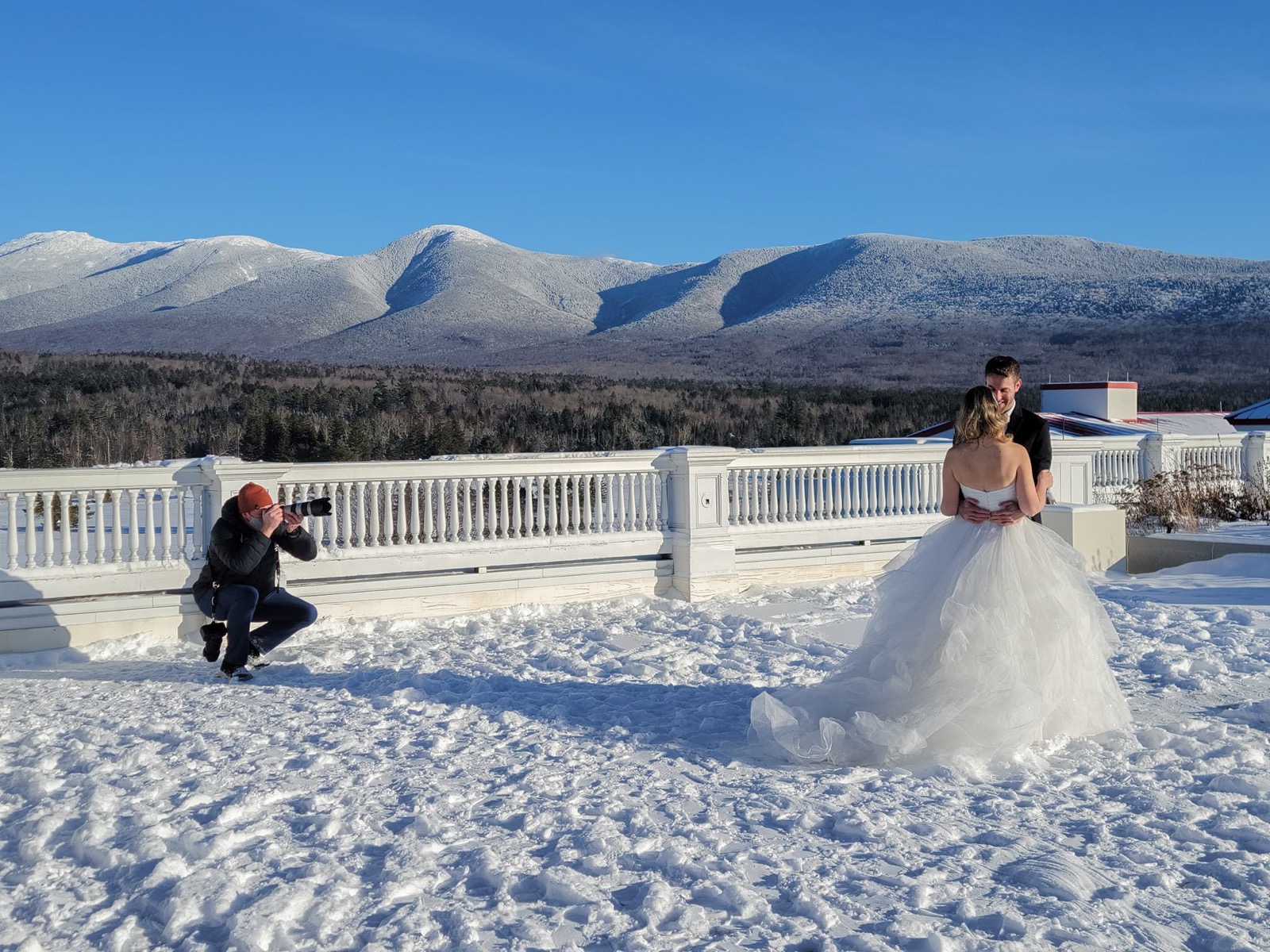 Eric crouched in the snow photographing a bride and groom on a terrace with the snow covered Presidential Range behind them