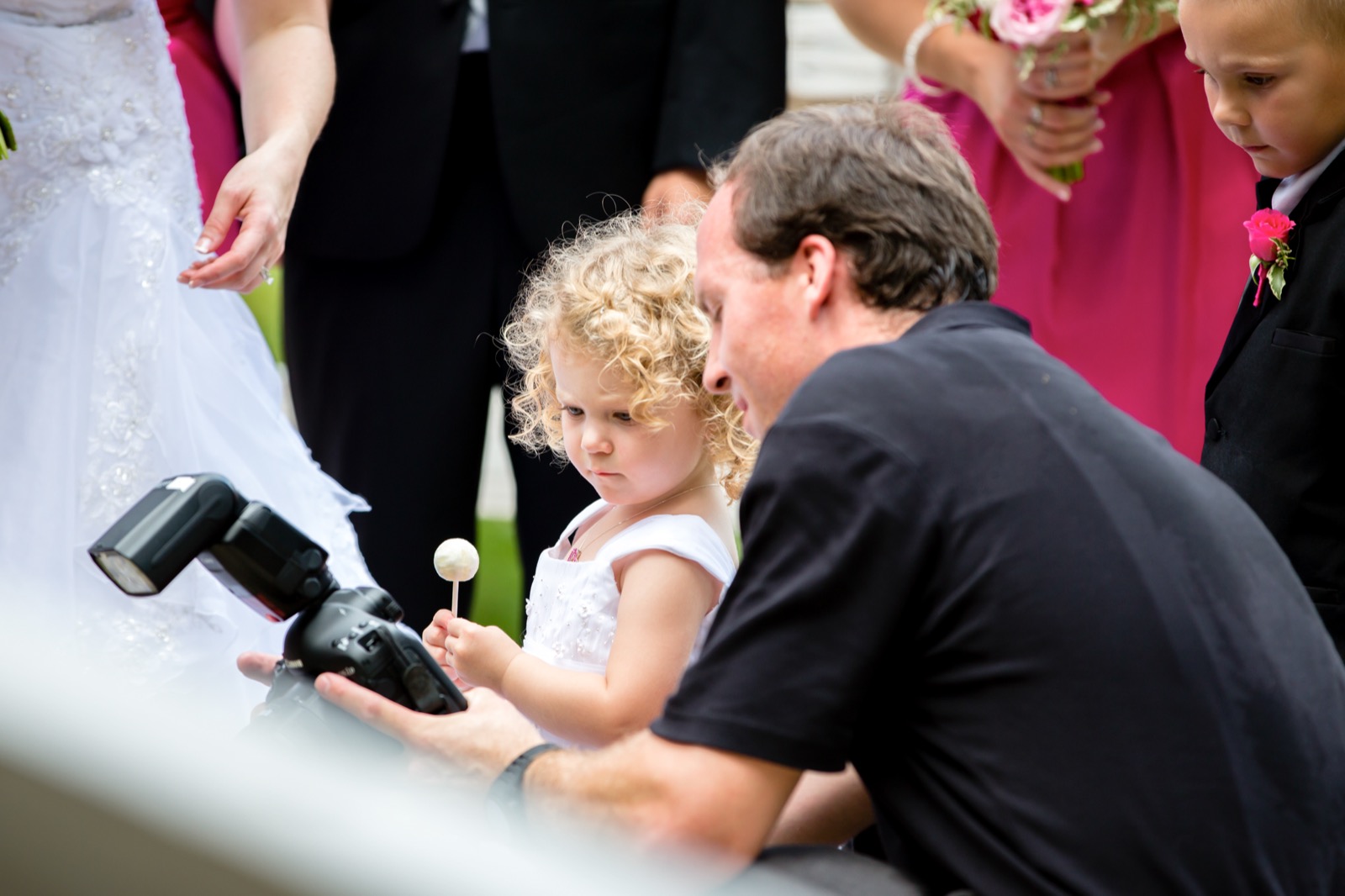 Eric kneeling beside a small flower girl and showing her the back of his camera during family portraits