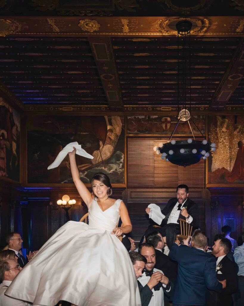 Bride and groom lifted on chairs during the hora as guests cheer in a wood-panelled ballroom