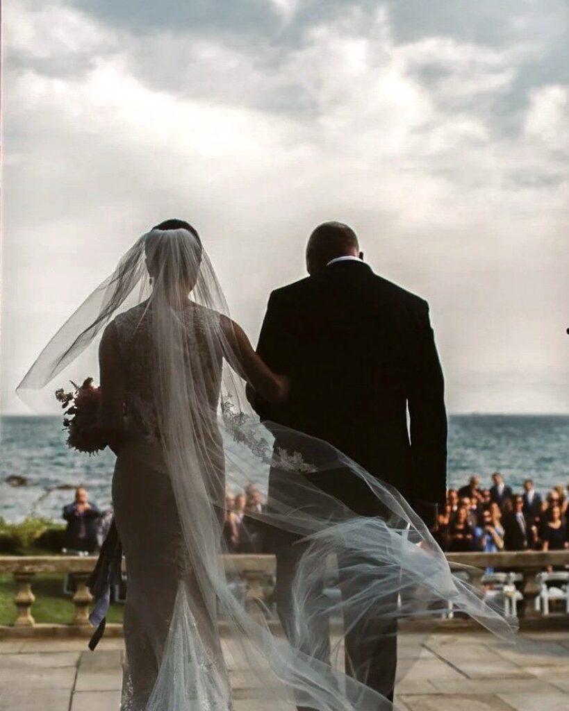 Bride and her father walk toward an oceanfront ceremony, her veil lifting in the wind above the water