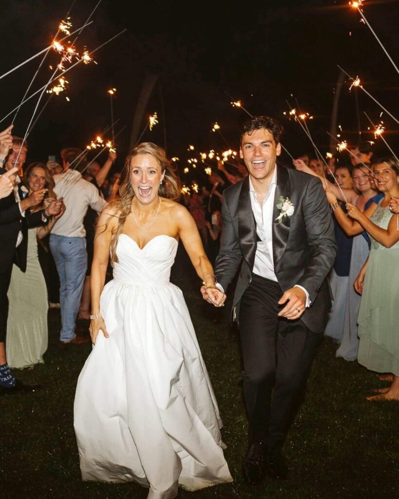Newlyweds run hand in hand through a sparkler send-off at night between two rows of guests
