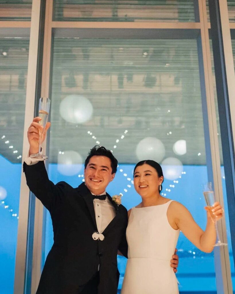Bride and groom raise a champagne toast in front of a blue-lit window wall hung with balloons