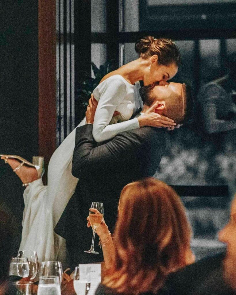 Groom lifts his bride into a kiss at the reception as a seated guest watches with a glass of champagne