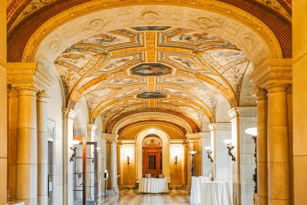 The vaulted marble hall at Aldrich Mansion with painted ceiling panels, stone columns and white-draped cocktail tables set for a wedding cocktail hour
