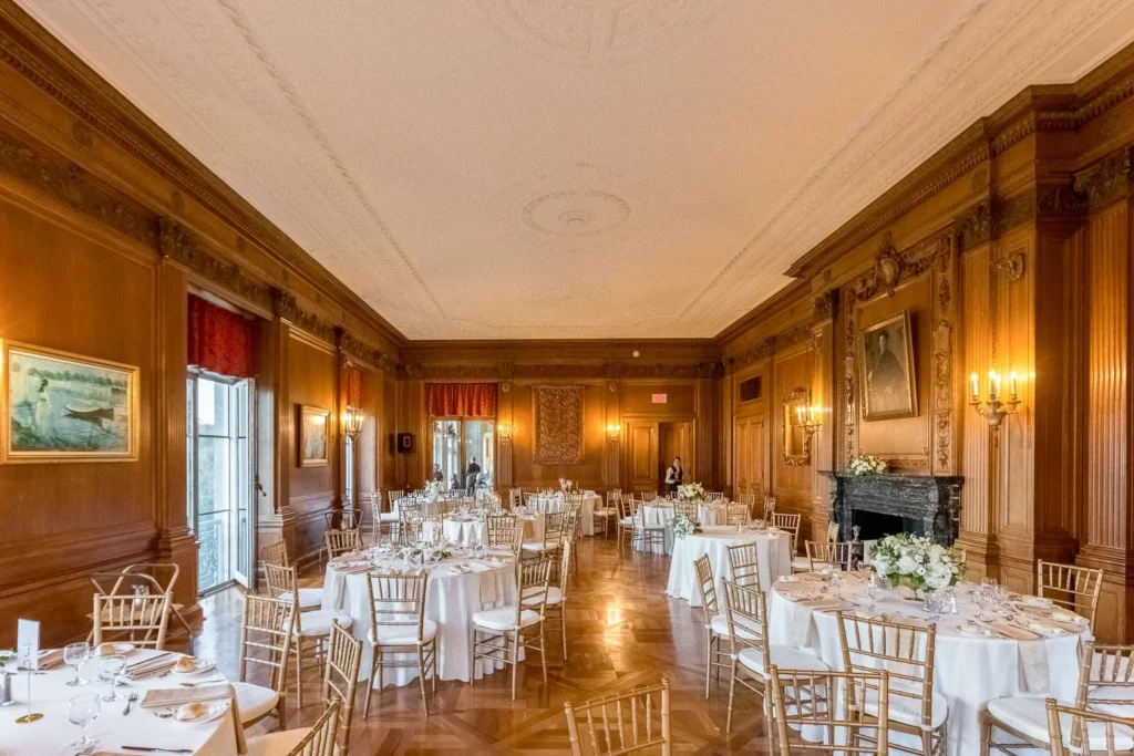 A wood-paneled dining room at Aldrich Mansion set for a wedding reception with round tables, white linens, gold Chiavari chairs and a carved fireplace