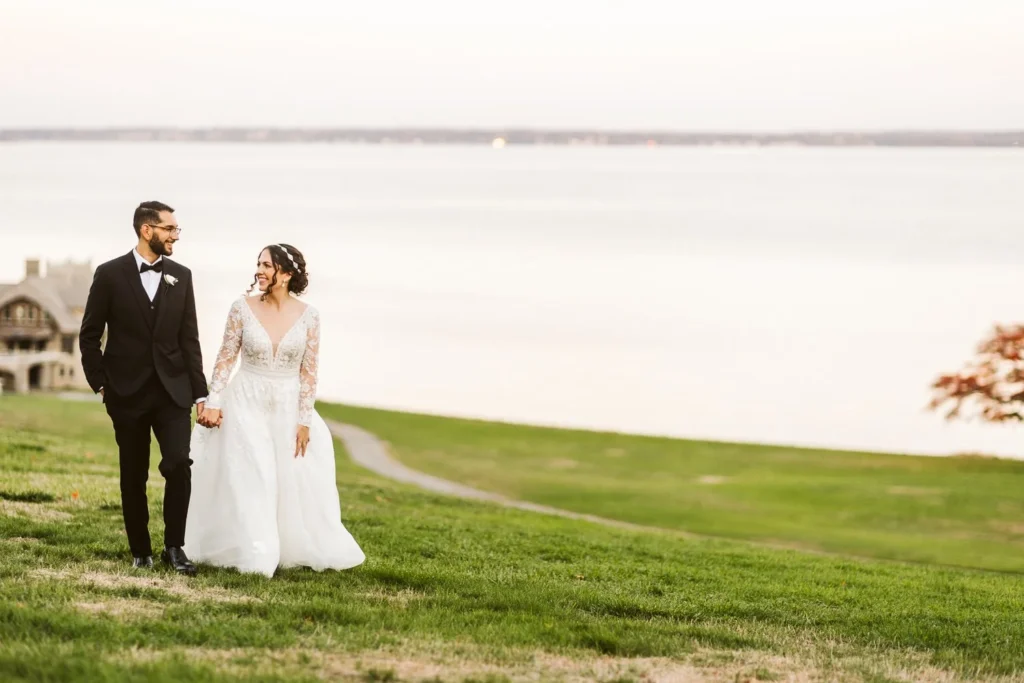 Bride and groom walk hand in hand across the lawn at Aldrich Mansion with Narragansett Bay flat and silver behind them