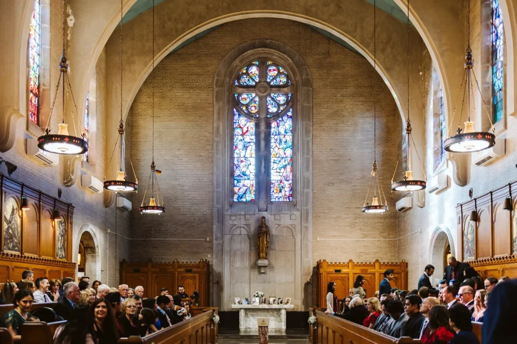 Guests fill the pews of the stone chapel at Aldrich Mansion during a Catholic wedding ceremony, tall stained-glass windows and iron pendant lights above the altar