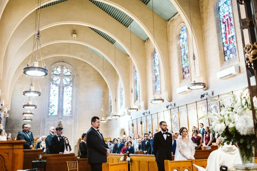 Bride and groom stand at the altar of the chapel at Aldrich Mansion with the groomsmen beside them, stained-glass windows and white florals along the pews