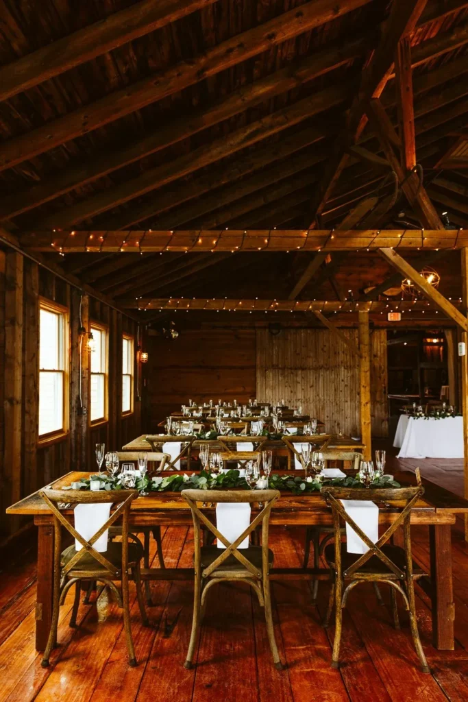 Inside the Barn at Bradstreet Farm set for a wedding with long handcrafted farm tables, wooden cross-back chairs, greenery and string lights under the timber beams