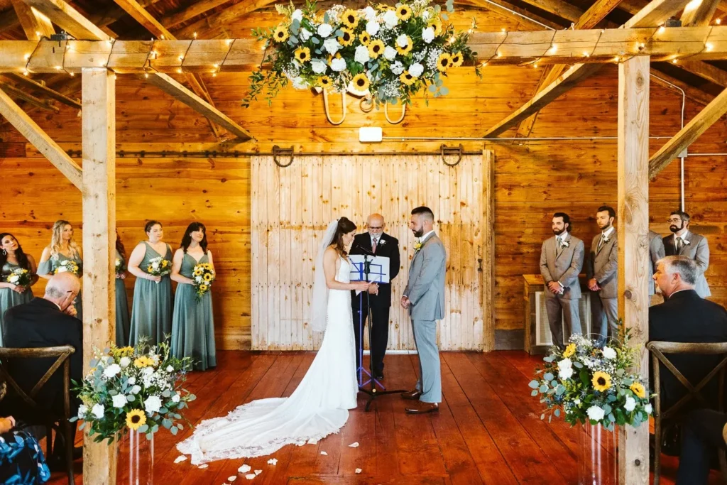 A wedding ceremony inside the Barn at Bradstreet Farm, the couple and officiant at the barn doors under a sunflower arrangement with the wedding party on either side