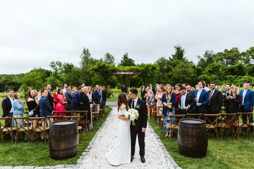 A bride and her father walk the white gravel aisle between whiskey barrels and cross-back chairs at an outdoor ceremony at the Barn at Bradstreet Farm as guests stand