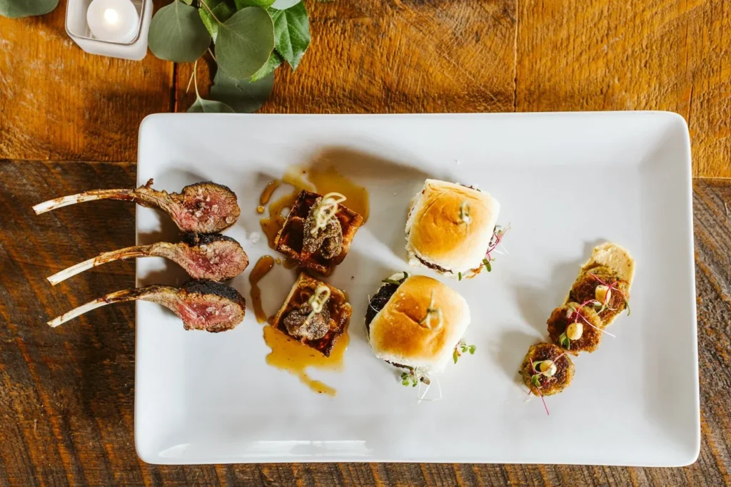 Lamb chops and mini sliders plated on a white tray on a wooden farm table at a Barn at Bradstreet Farm wedding