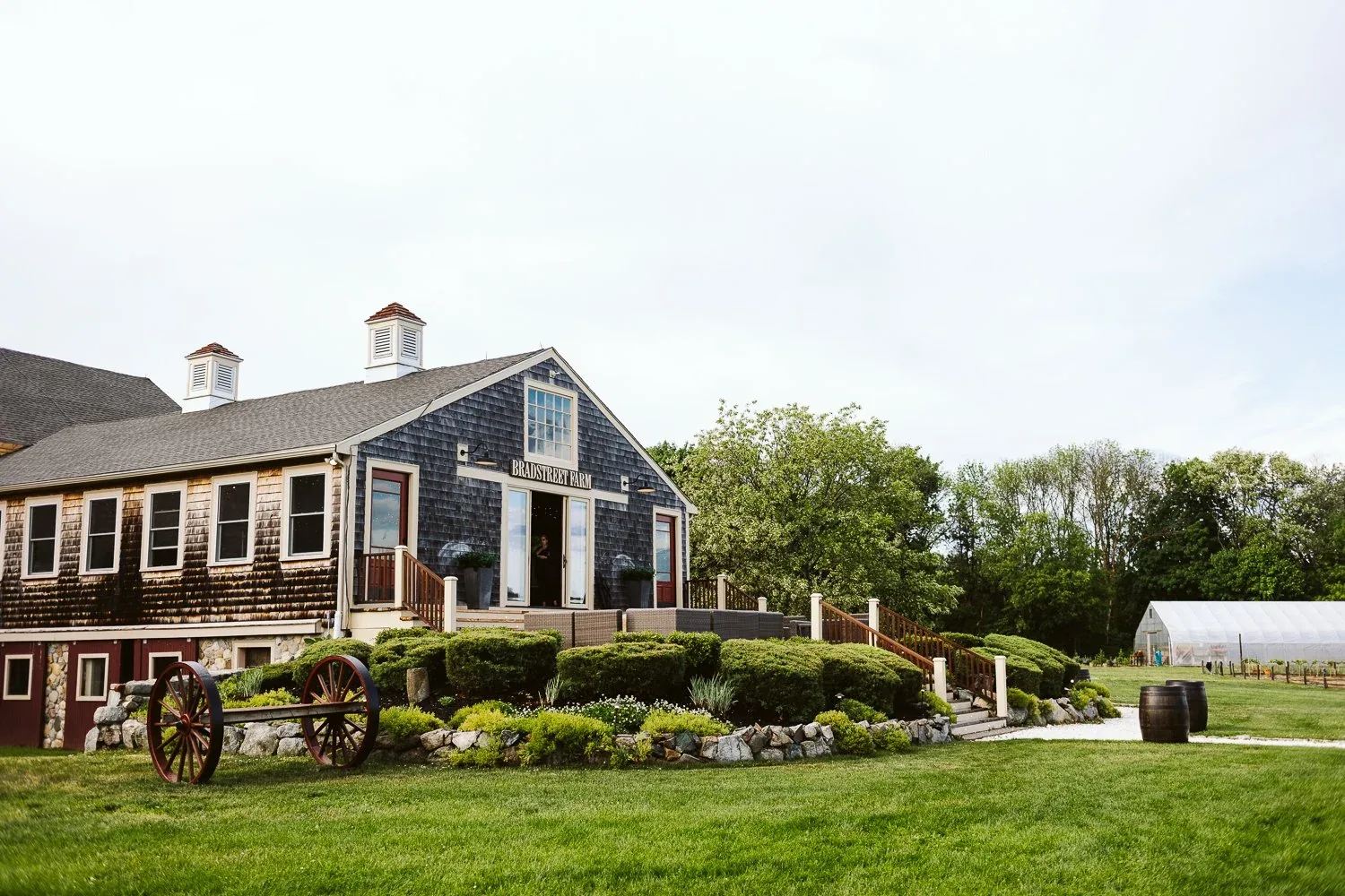 The Barn at Bradstreet Farm Wedding 1 The restored 18th-century grey-shingled barn at Bradstreet Farm in Rowley, MA, with a cupola, an antique wagon wheel and stone-edged garden beds on the lawn