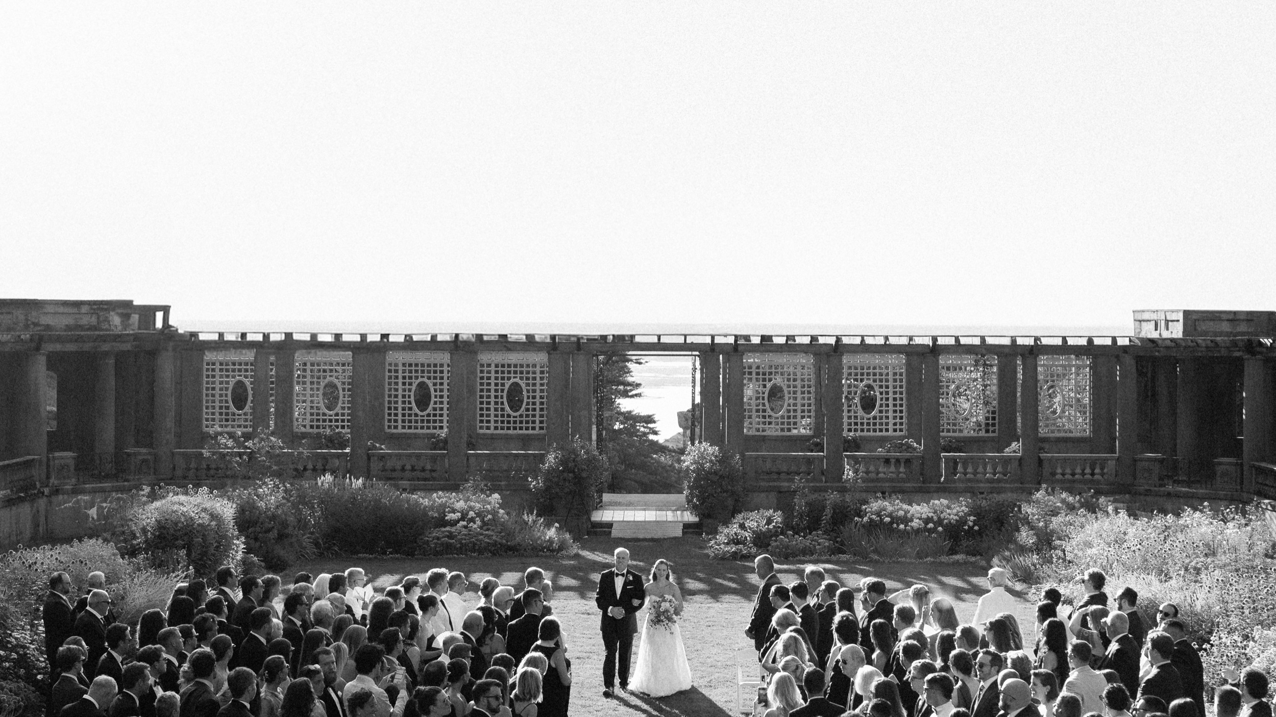 A bride and her father walk the aisle at a Crane Estate wedding ceremony in Ipswich, the pergola and the sea behind them
