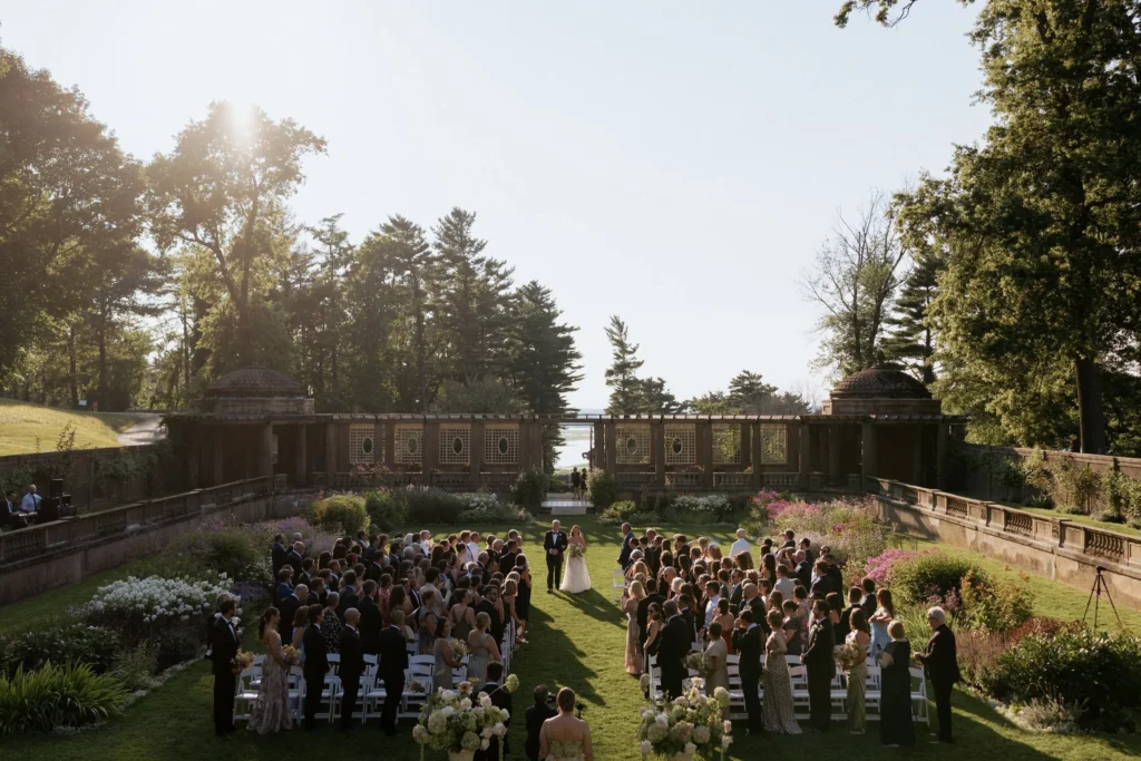 Wide view of a wedding ceremony in the sunken Italian Garden at the Crane Estate, guests seated on the lawn between the stone walls with the sun low over the trees