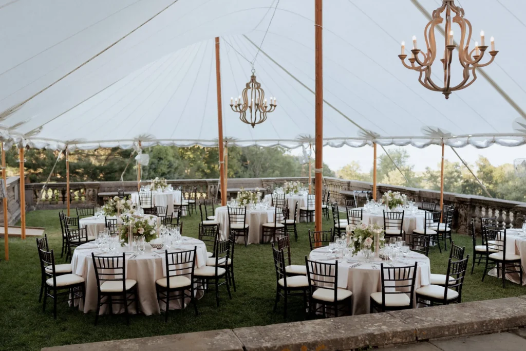 A sailcloth tent with chandeliers set for dinner on the east terrace of the Great House at the Crane Estate, round tables with black chairs and the lawn beyond