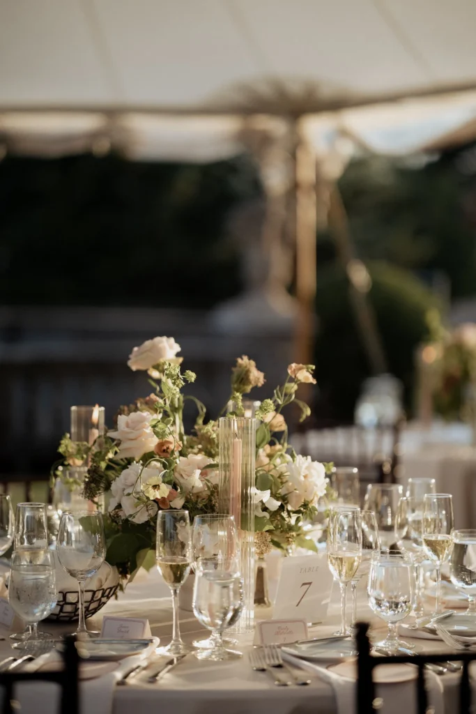 A dinner table under the sailcloth tent at the Crane Estate with garden roses, taper candles, glassware and a table number card