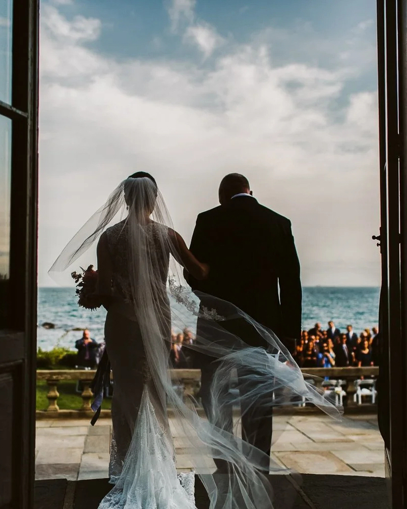A bride and her father step through the doorway toward a seaside ceremony in Sarasota, her veil lifting in the wind