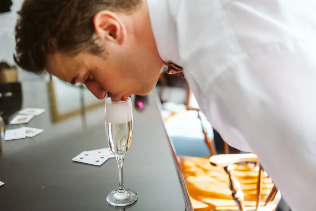Groom bends to sip from a full champagne flute on a bar with playing cards before his Hill-Stead Museum wedding