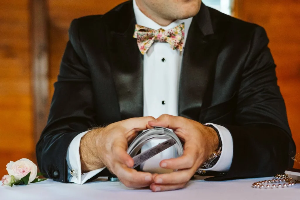 Groom in a black tuxedo and floral bow tie holds a silver flask with a rose and pearls on the table before his Hill-Stead wedding