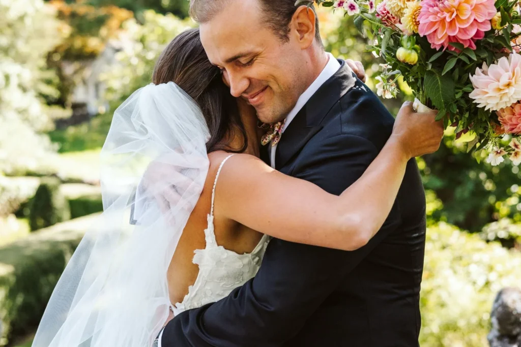 Bride and groom embrace during their first look on the pathway above the Sunken Garden at Hill-Stead Museum, her bouquet of dahlias over his shoulder