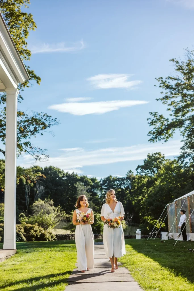 Two bridesmaids in white walk the path beside the veranda toward the West Lawn ceremony at Hill-Stead Museum