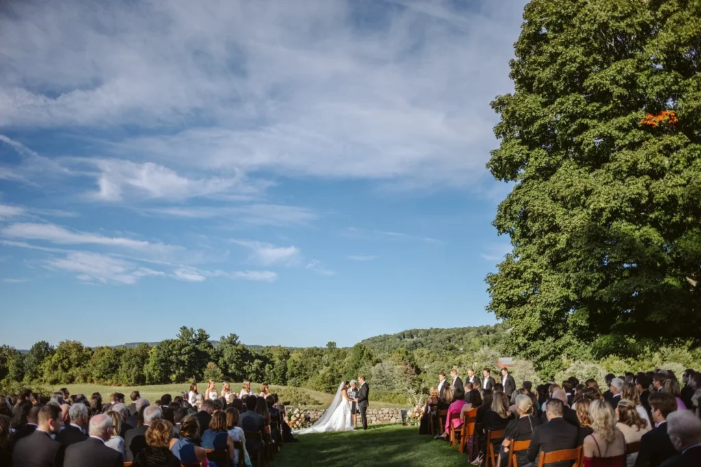 Wide view of the West Lawn ceremony at Hill-Stead Museum from behind the guests, with a big blue sky and the wooded hills beyond the meadow
