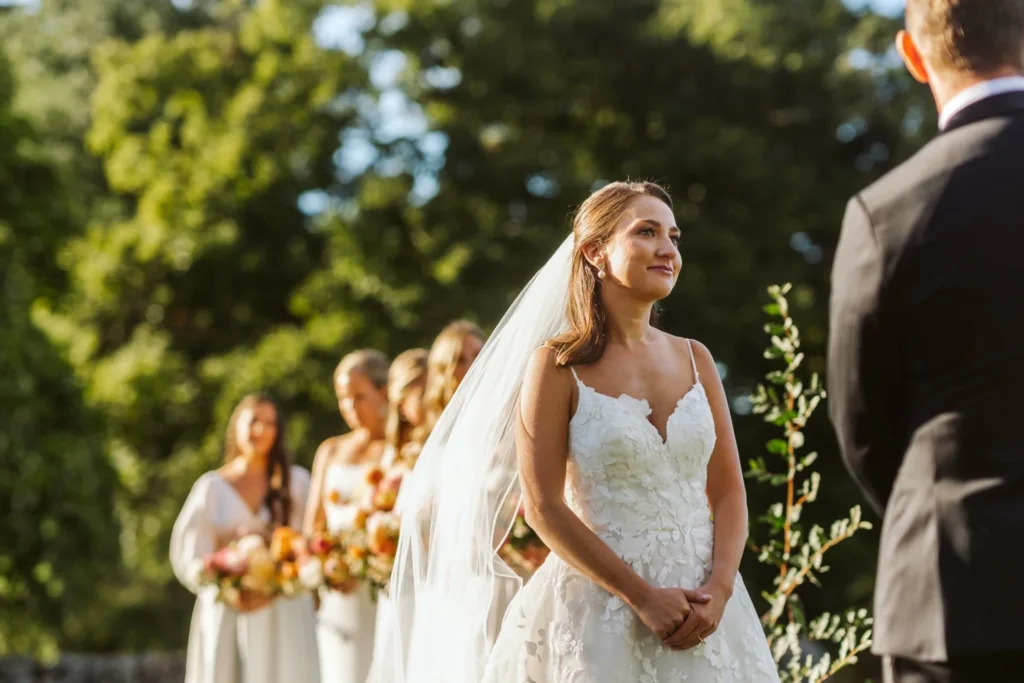Bride in a lace gown and veil listens at the altar with her bridesmaids behind her at Hill-Stead Museum