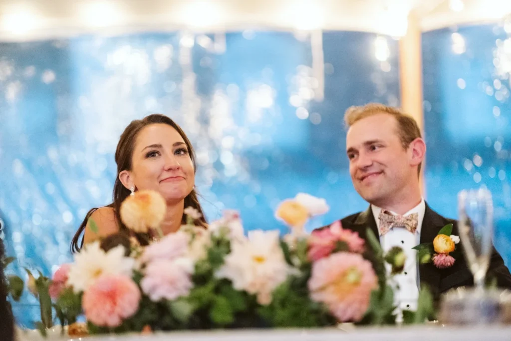 Bride and groom smile at the head table behind peach and pink florals during toasts under the tent at Hill-Stead Museum