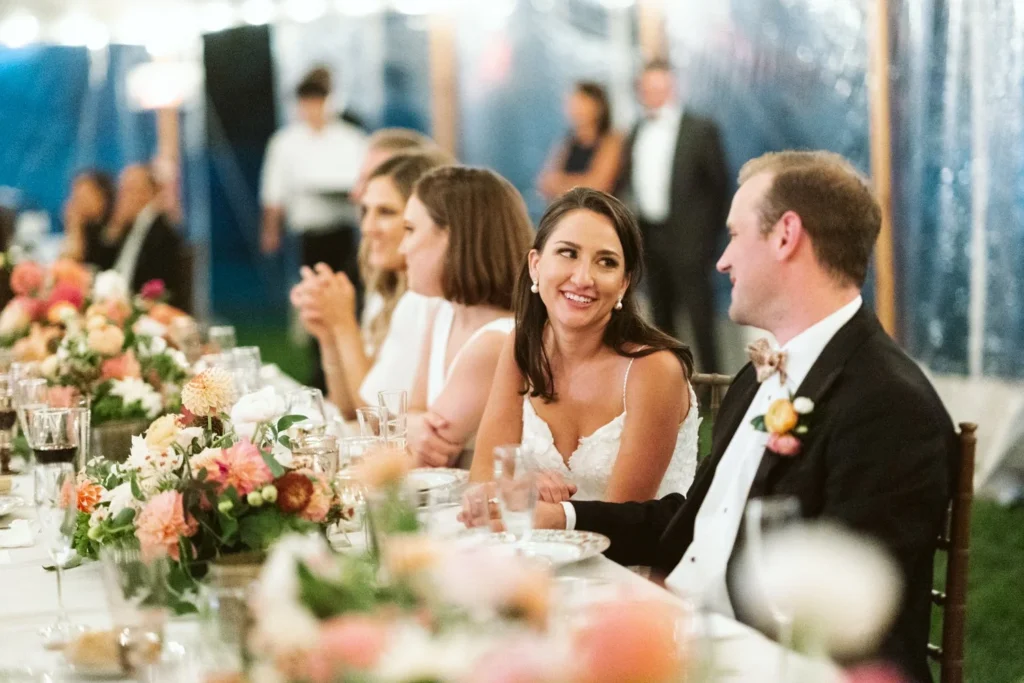 Bride and groom seated at the head table under the tent at Hill-Stead Museum with peach and pink dahlia arrangements in front of them