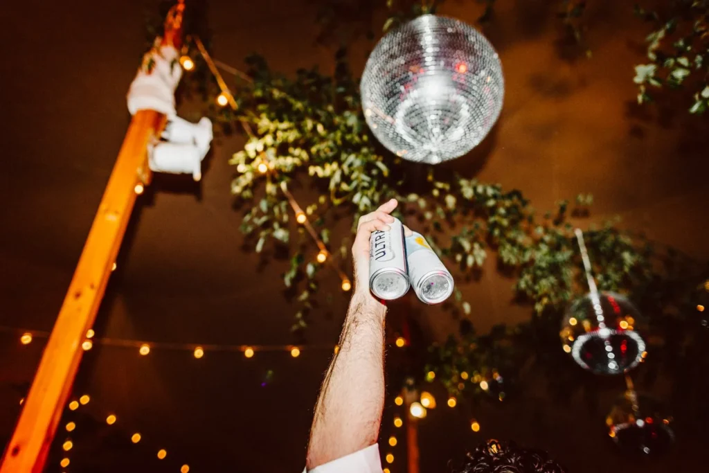 Hands raised with cans toward a disco ball and hanging greenery under the tent at Hill-Stead Museum