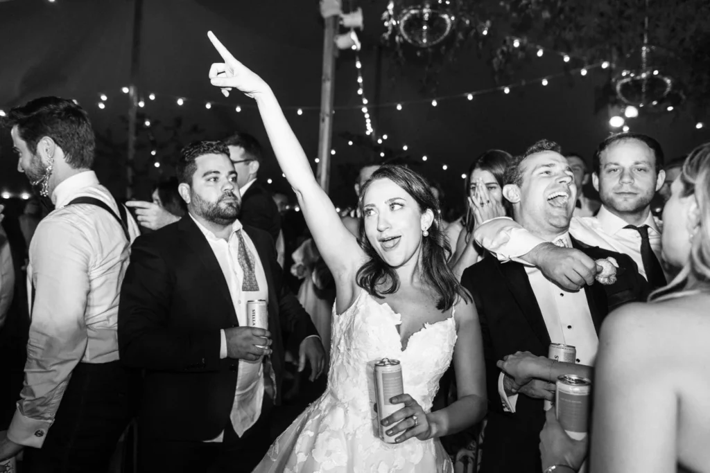 Black and white photo of the bride pointing skyward in a crowd of dancing guests under string lights at Hill-Stead Museum