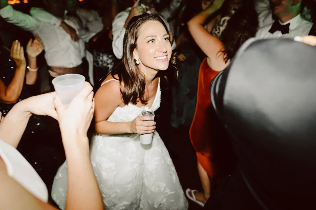 Bride with a drink in hand smiles through the crowd on the dance floor late at night at Hill-Stead Museum