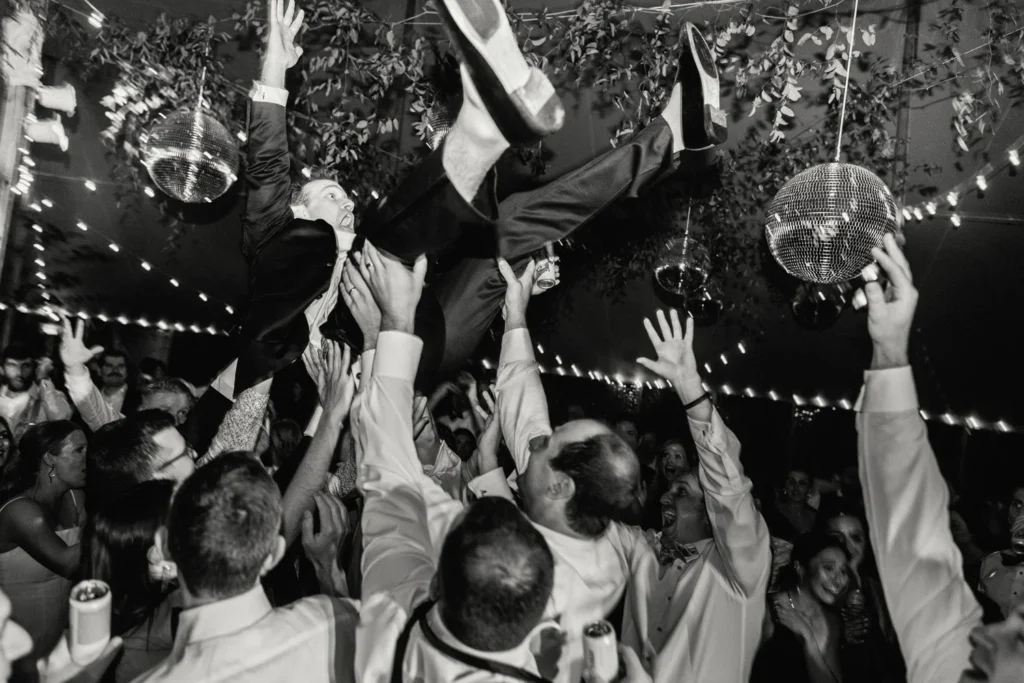 Black and white photo of guests lifting a man above the crowd under disco balls at Hill-Stead Museum