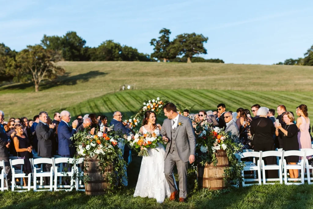 Guests seated on the lawn at The Barn at Gibbet Hill watch a wedding ceremony at a flower-trimmed birch arbor with the green rolling hills of the farm and a line of trees behind