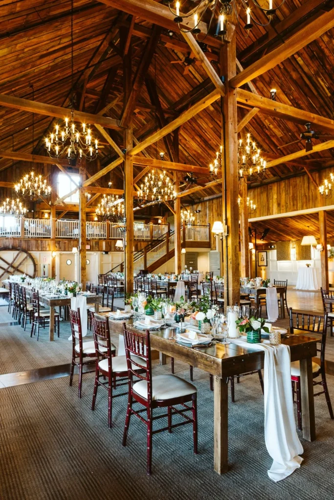 The post-and-beam interior of The Barn at Gibbet Hill set for a wedding reception with long wooden tables, wooden chairs, greenery and chandeliers hanging from the rafters