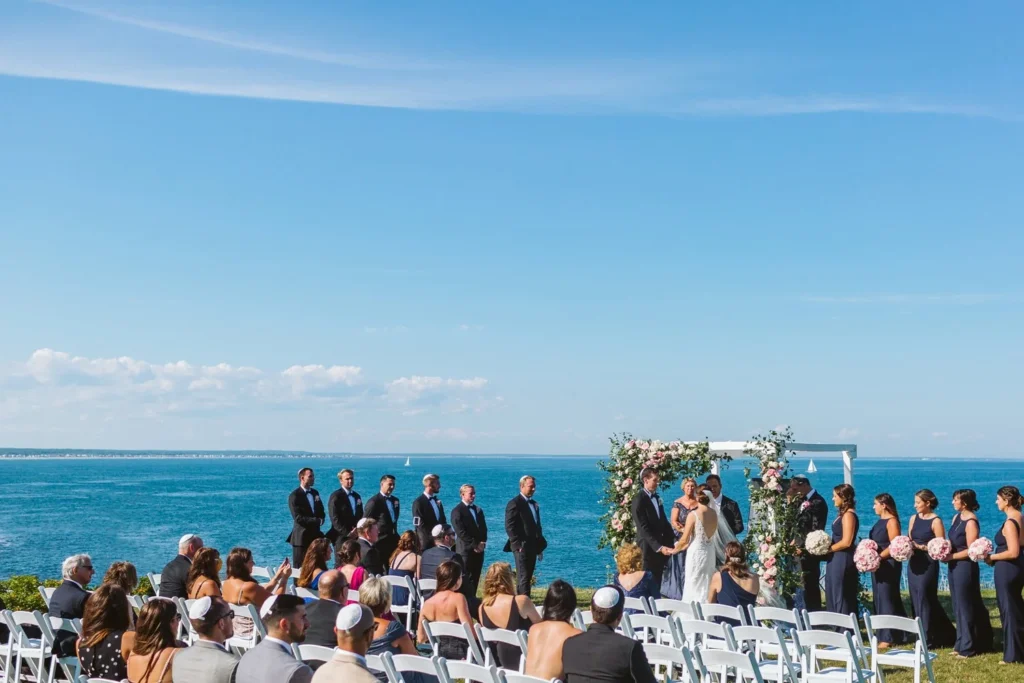 A wedding ceremony on the North Point Lawn at Cliff House Maine with the wedding party at a floral arch and the Atlantic Ocean behind them