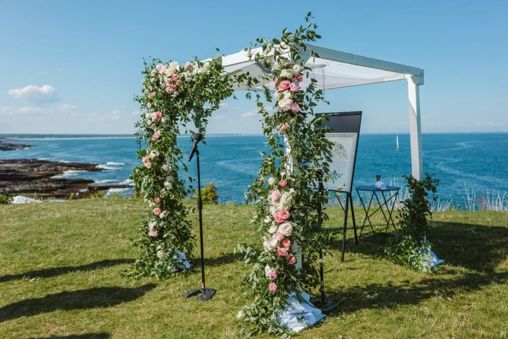 A white arch covered in pink and white roses and greenery on the North Point Lawn at Cliff House Maine before the ceremony, with the rocky coast and ocean behind