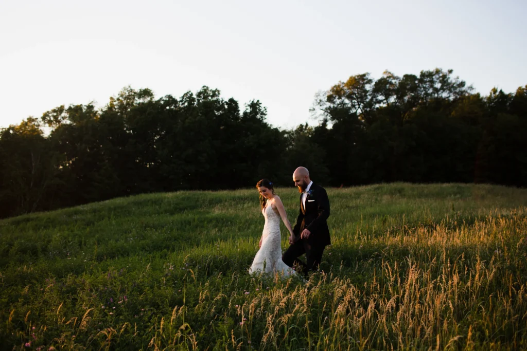 Bride and groom walk hand in hand through a tall-grass meadow at Tower Hill Botanic Garden in the last light of the evening