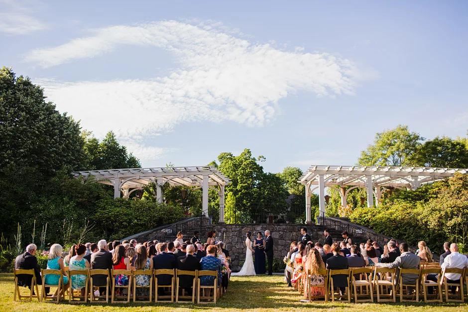 Wedding guests seated on the lawn at Tower Hill Botanic Garden with the white pergolas and stone staircase behind the ceremony