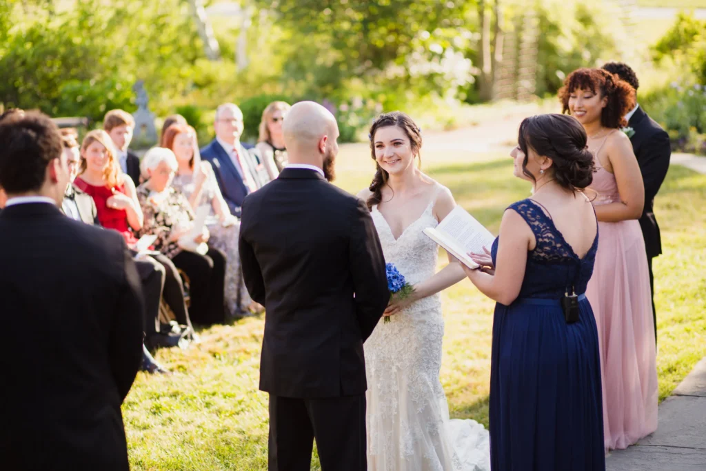 Bride and groom stand with their officiant in the Secret Garden at Tower Hill Botanic Garden while guests watch from the grass