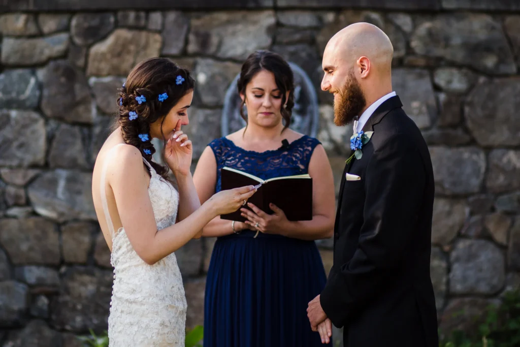 Bride reads her vows beside the officiant and groom in front of the stone wall of the Secret Garden at Tower Hill Botanic Garden