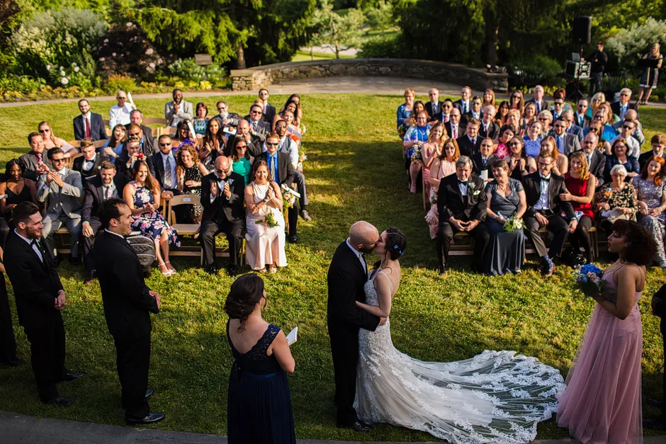 Bride and groom kiss at the end of their Secret Garden ceremony at Tower Hill Botanic Garden as guests seated on the grass applaud